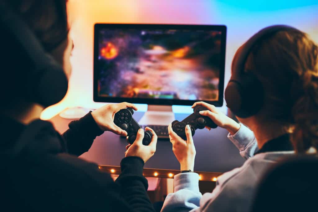 Friends playing video game at home. Gamers holding gamepads sitting at front of screen. Streamers girl and boy playing online in dark room lit by neon lights. Competition and having fun Friends playing video game at home. Gamers holding gamepads sitting at front of screen. Streamers girl and boy playing online in dark room lit by neon lights. Competition and having fun