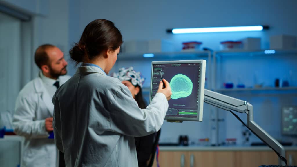 Researcher looking at monitor analyzing brain scan while coworker discussing with patient in background about side effects, mind functions, nervous system, tomography scan working in laboratory