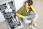 happy woman sitting near the dishwasher with clean dishes on the kitchen at home. Easy house work with kitchen appliances concept