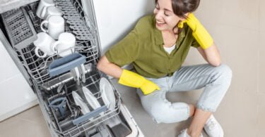 happy woman sitting near the dishwasher with clean dishes on the kitchen at home. Easy house work with kitchen appliances concept